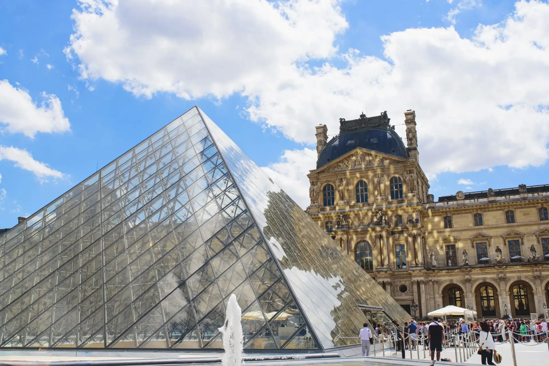 View of the Louvre Museum, the world's largest art museum and a historic monument in Paris, France, on a sunny day. People are lining up outside, with a fountain visible in the foreground.