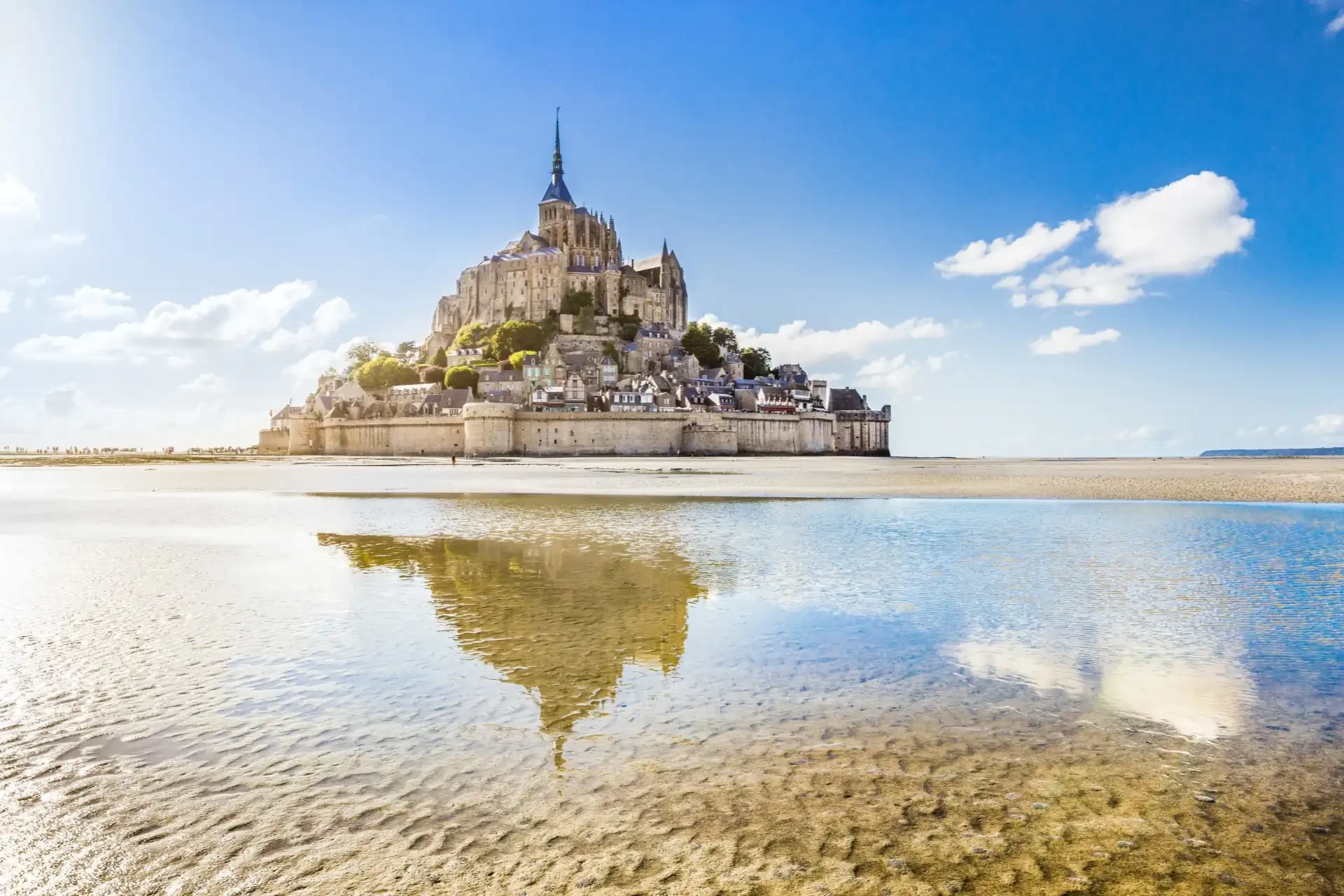 Mont Saint-Michel reflected in the water, with blue skies and some clouds visible. The surroundings are clear, highlighting the picturesque scene.