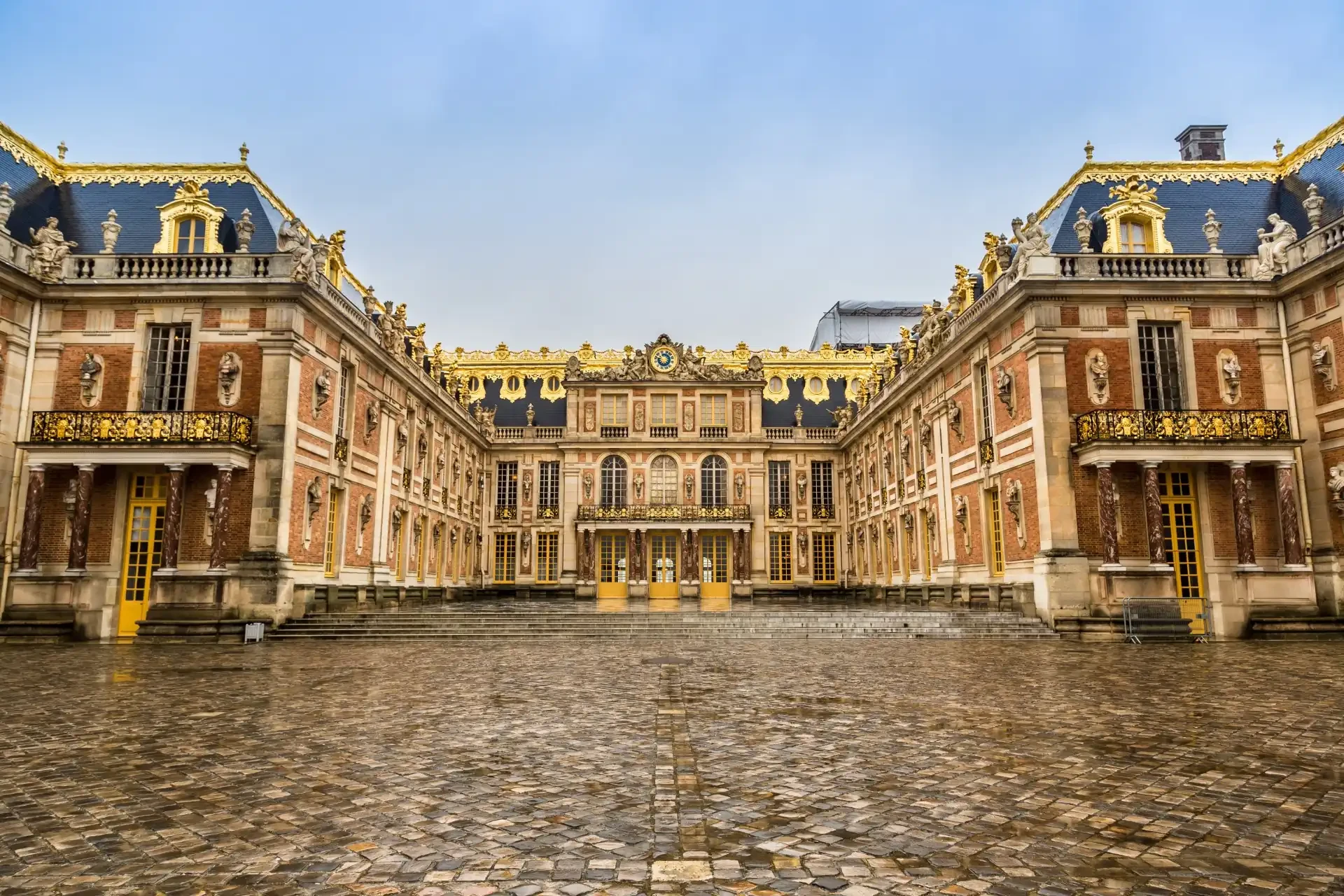 Exterior view of the Palace of Versailles with cobblestones leading up to the grand building. The palace's intricate architecture is prominently displayed.