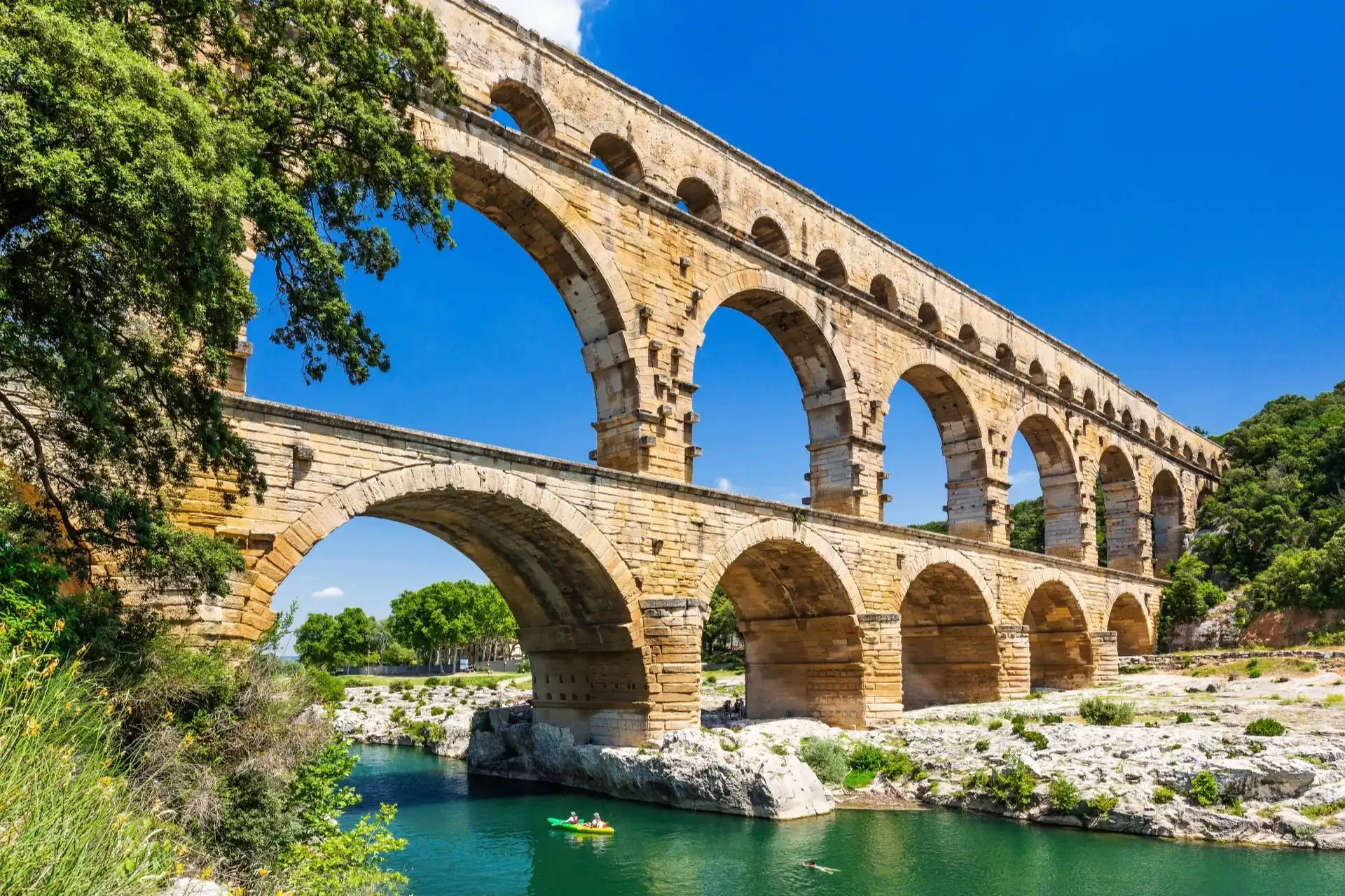 The Pont du Gard aqueduct with kayakers enjoying the river below. Green trees and plants surround the scene, enhancing its beauty.