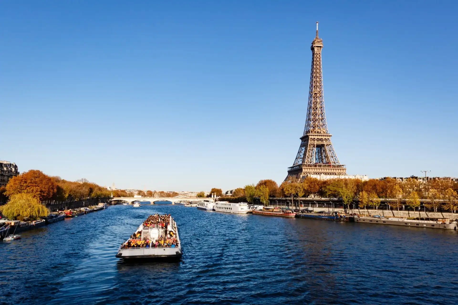 People in a boat approaching the Eiffel Tower on the Seine River. Autumn foliage is visible in the background.