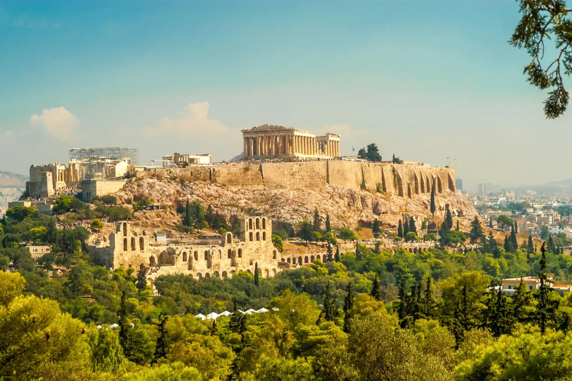 The Acropolis of Athens against a backdrop of a blue cloudy sky, with green foliage in the foreground.