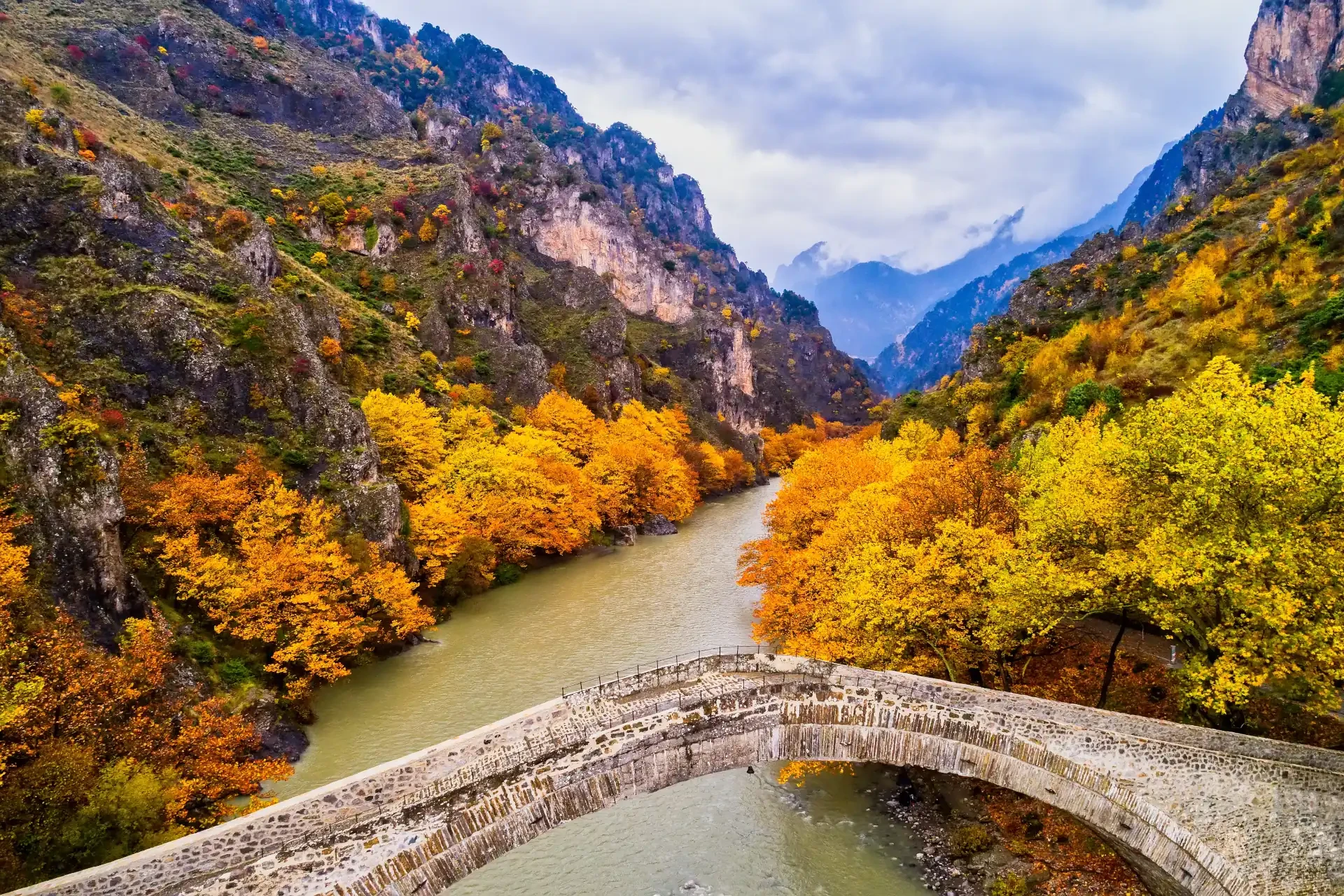 An aerial view of Konitsa old bridge and Aoos River on an autumn day in Greece. Yellow, orange, and brown leaves cover the ground under a cloudy sky.