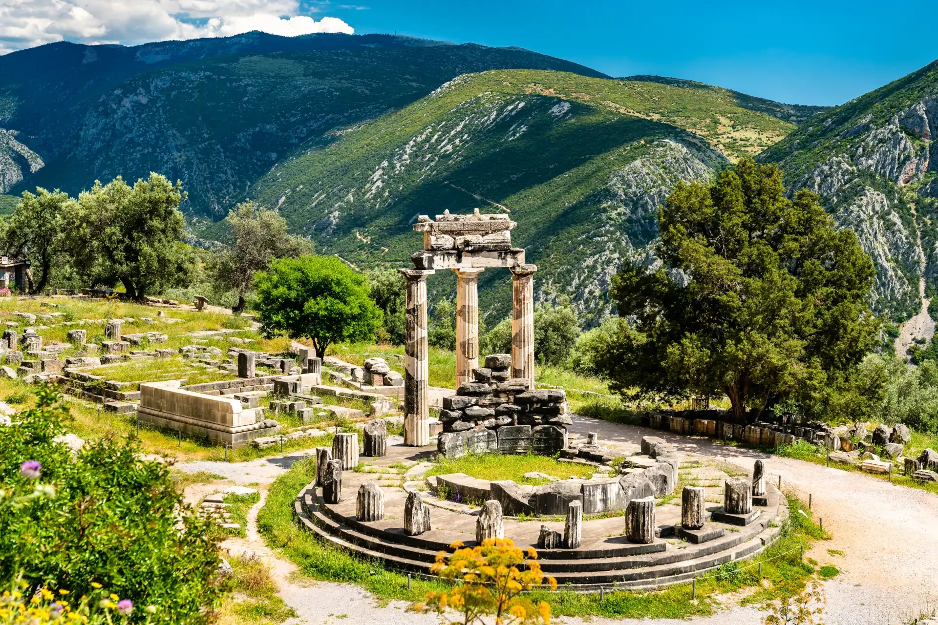 Temple of Athena Pronaia amidst the ruins of Delphi in Greece. Grass grows among ancient stones with a mountainous backdrop and blooming flowers.