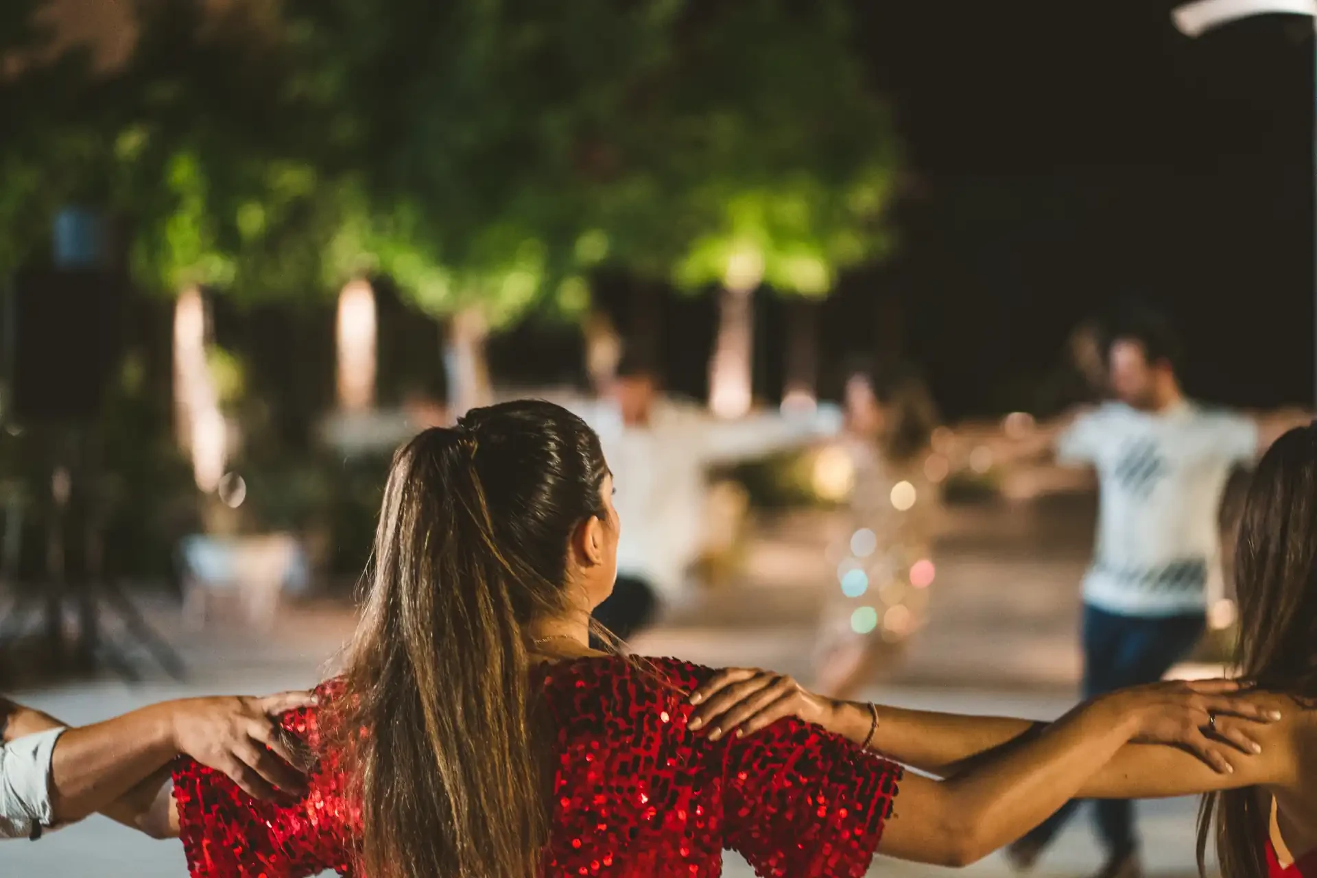 A traditional Greek dance performance with participants holding shoulders in a circle. A person in the foreground with dark hair wears a red sequinned dress.