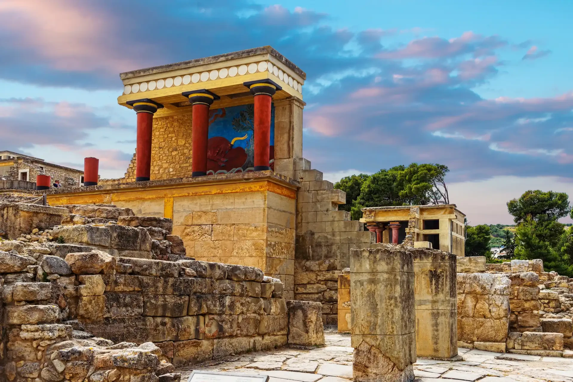 Knossos archaeological site in Crete, Greece. Distinctive red pillars and remnants of ancient paintings on structures, with a pink and blue cloudy sky and trees in the background.