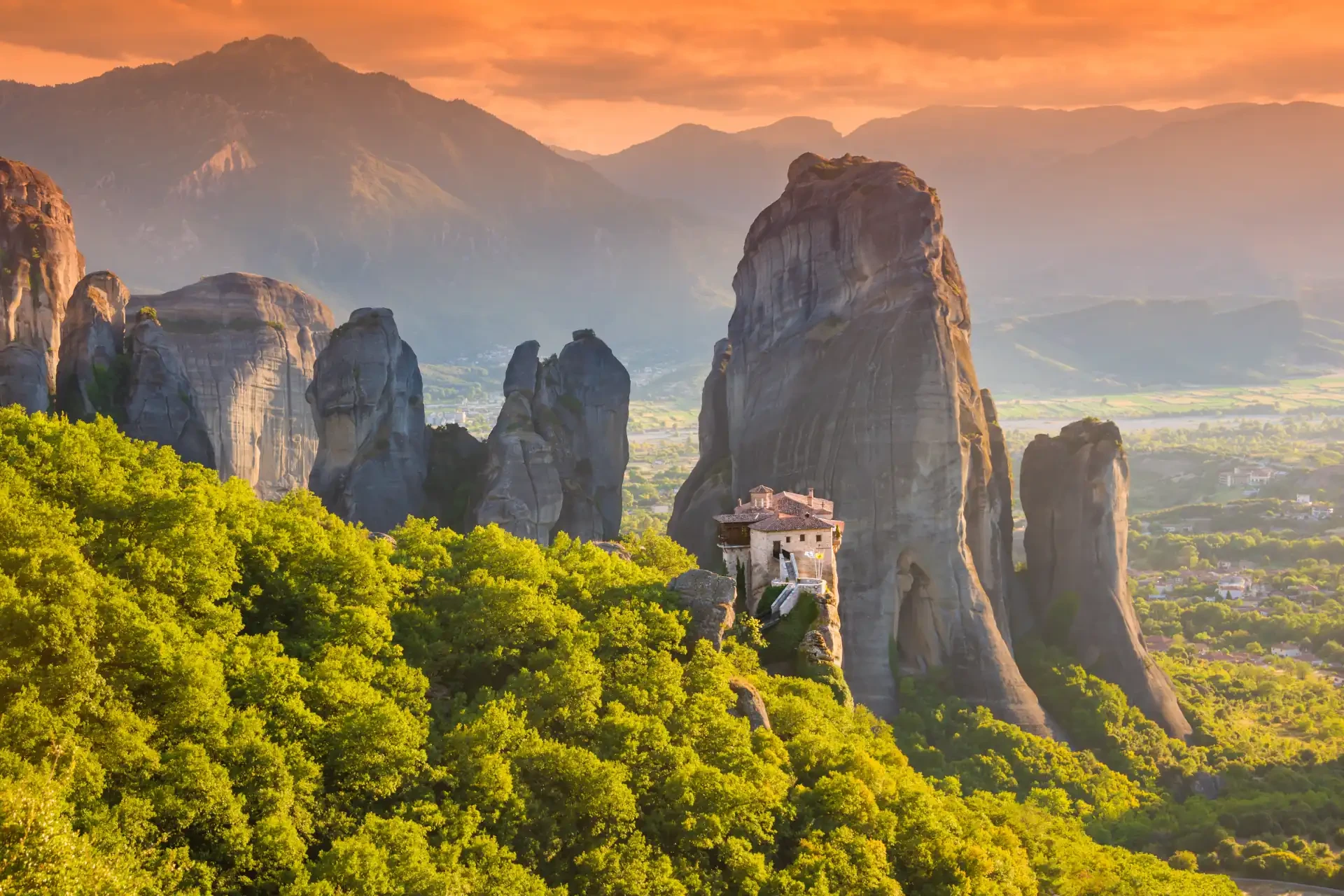 One of the Meteora monasteries built on rocky cliffs, set against a sunset sky with green foliage in the foreground and a mountainous backdrop.