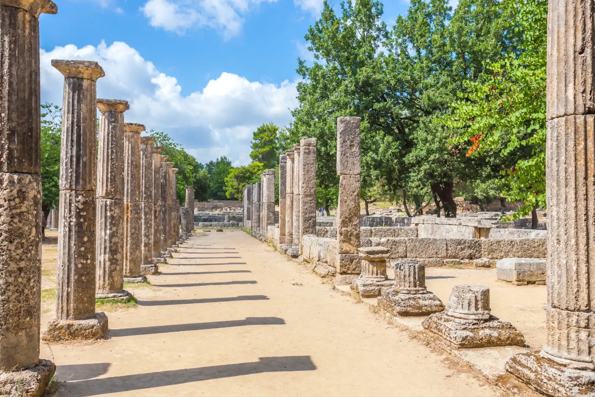 Ruins of Ancient Olympia in Peloponnese, Greece, under a blue cloudy sky. Surrounding green trees and shadows cast by ancient pillars on the pathway.