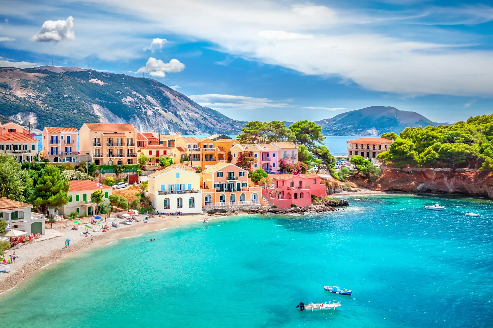 Assos village in Kefalonia, Greece, with colourful houses along turquoise waters. A boat floats in the foreground, with people on a sandy beach in the background.