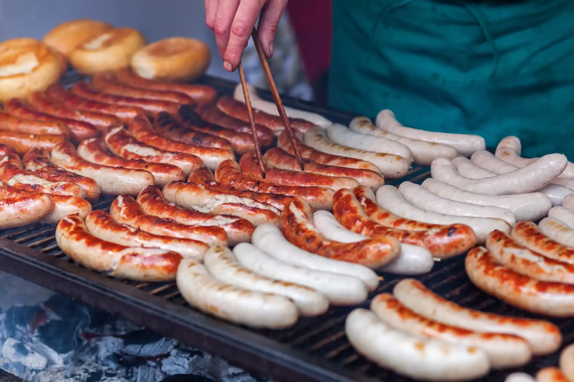 Rows of bratwurst sausages being grilled. A person uses tongs to rotate them.