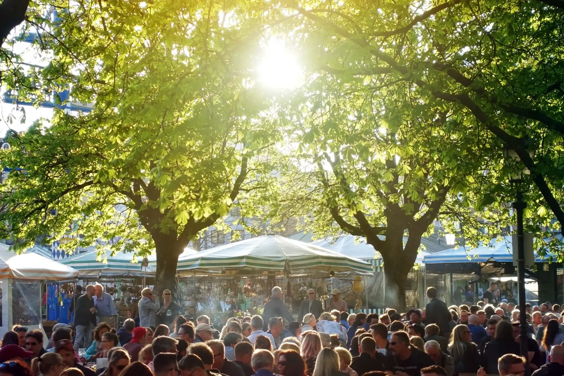 A crowd of people standing in front of a beer tent in a Munich beer garden. The sun shines through the trees at the scene.