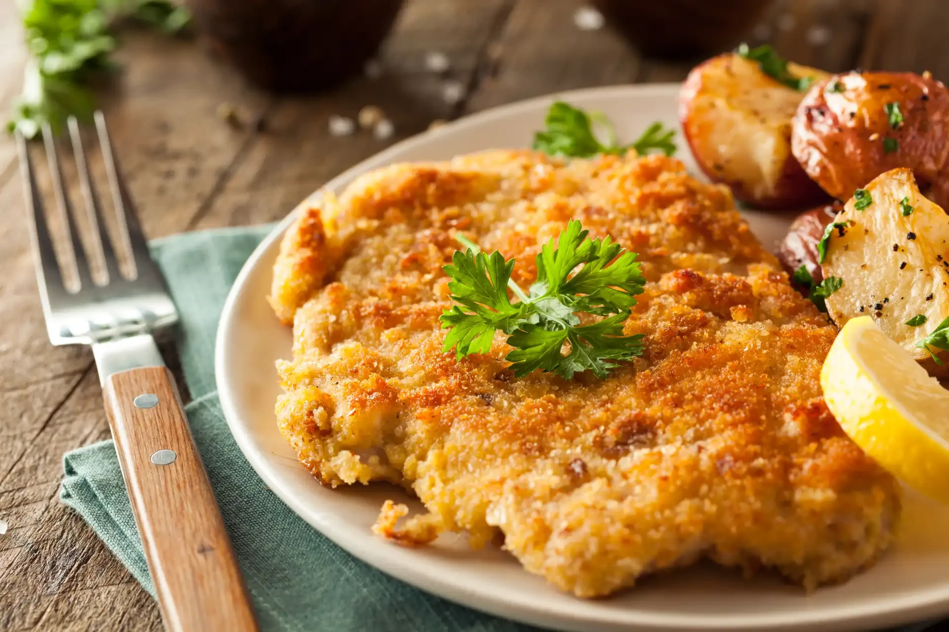 Homemade breaded German Wiener Schnitzel, served on a plate with a crispy golden-brown coating. Parsley and lemon are served as garnishes alongside potatoes.