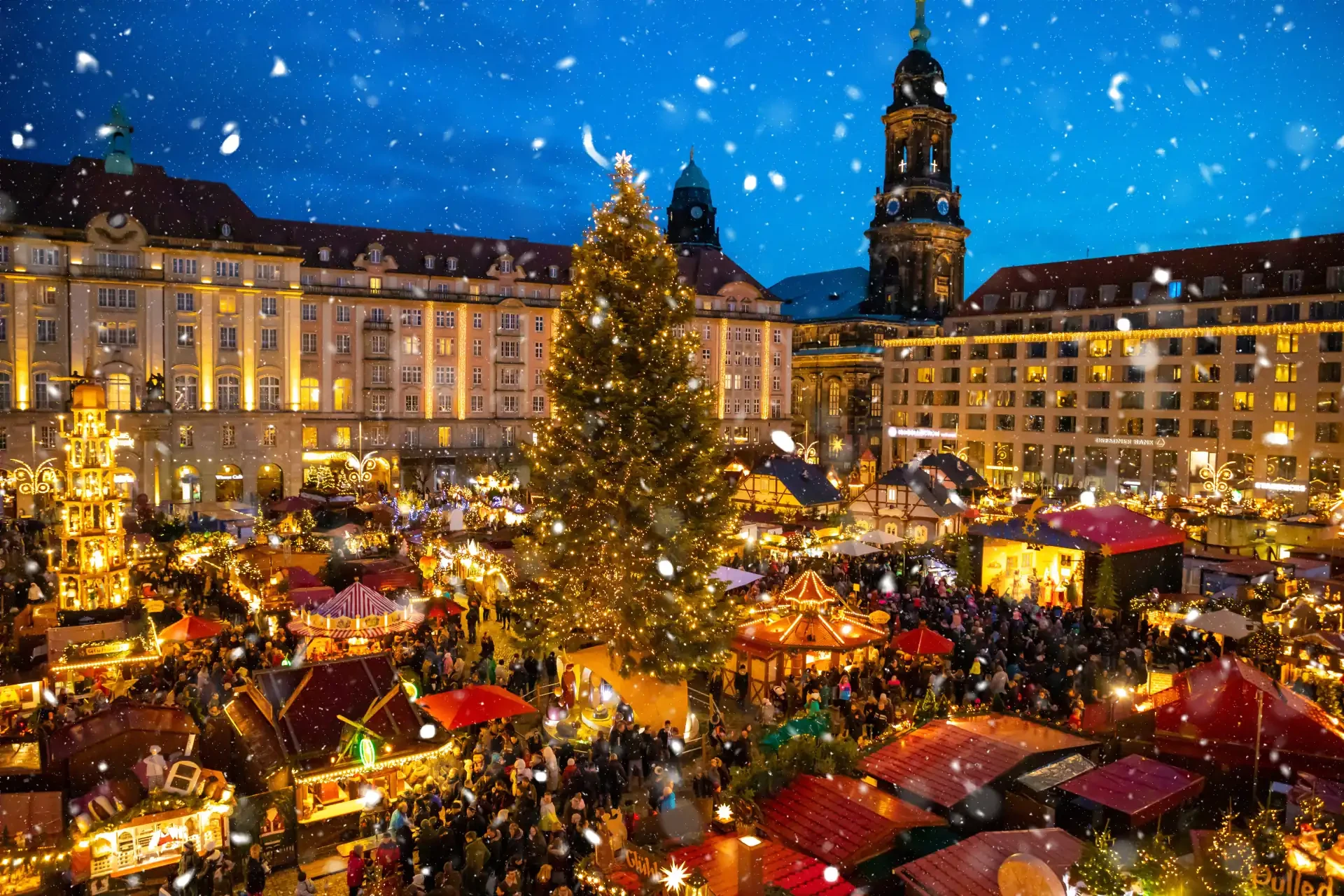 People visiting the Christmas Market Striezelmarkt in Dresden, Germany, with festive stalls and holiday decorations. Flakes of snow can be seen falling from the sky.