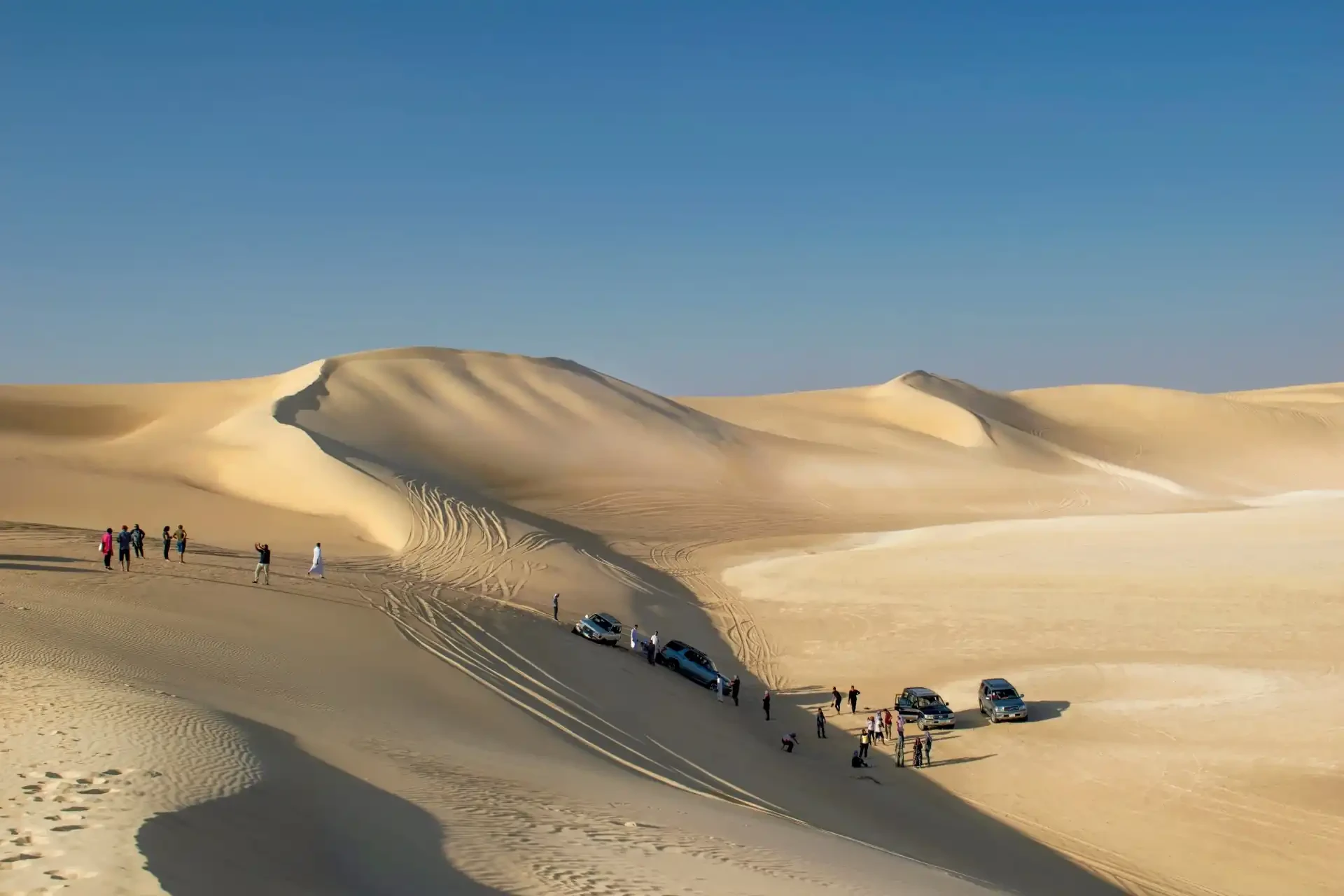 Serene desert landscape with pale sand, people, and parked cars under a clear blue sky in Siwa Oasis.