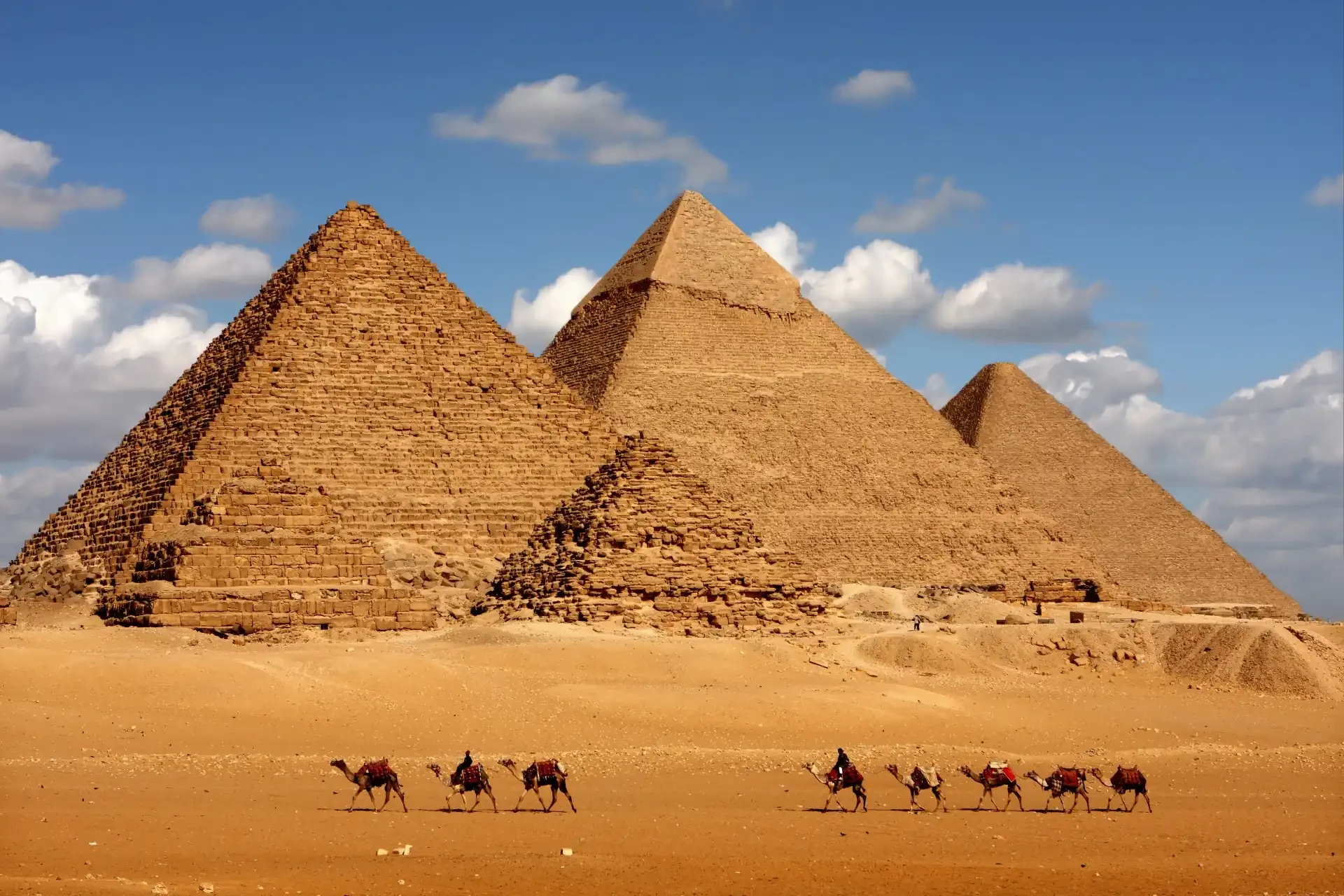 Majestic pyramids against a cloudy blue sky. Camels can be seen in the foreground.