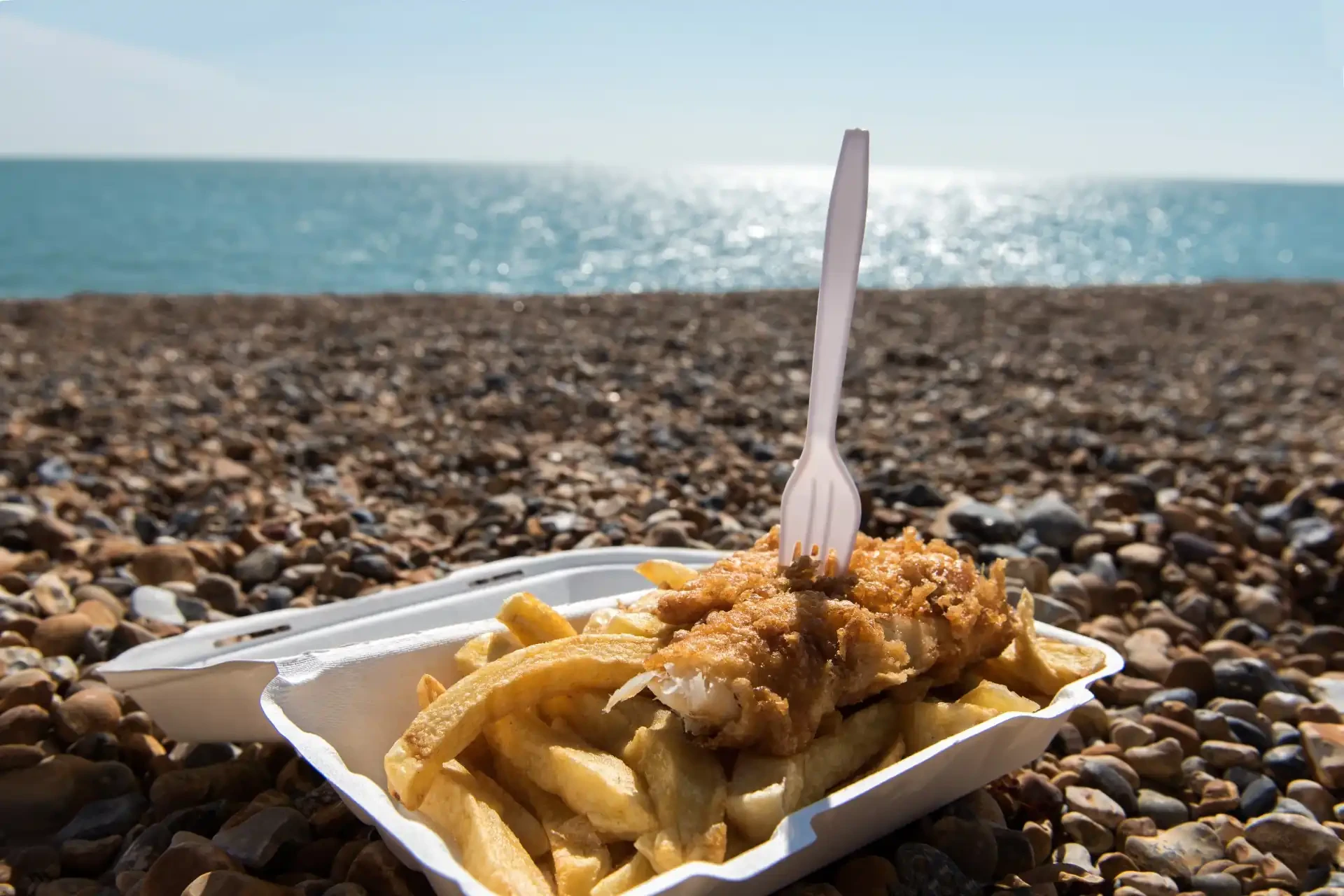 A seaside scene featuring a classic dish of fish and chips served in a polystyrene tray.