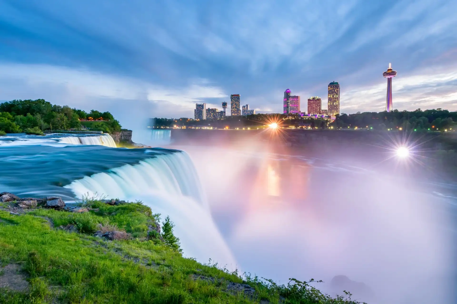Niagara Falls in Ontario, a world-famous natural wonder with powerful cascading waterfalls. The Toronto skyline can be seen in the background, featuring the iconic CN Tower.