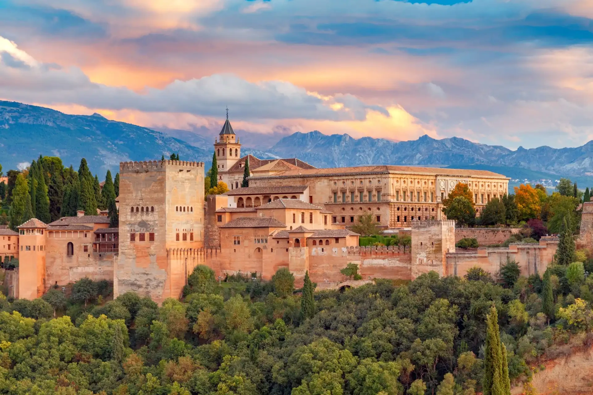The Alhambra fortress and palace complex in Granada, Spain, is depicted during sunset, with mountains in the background and an orange and blue-grey sky casting a warm glow over the scene.