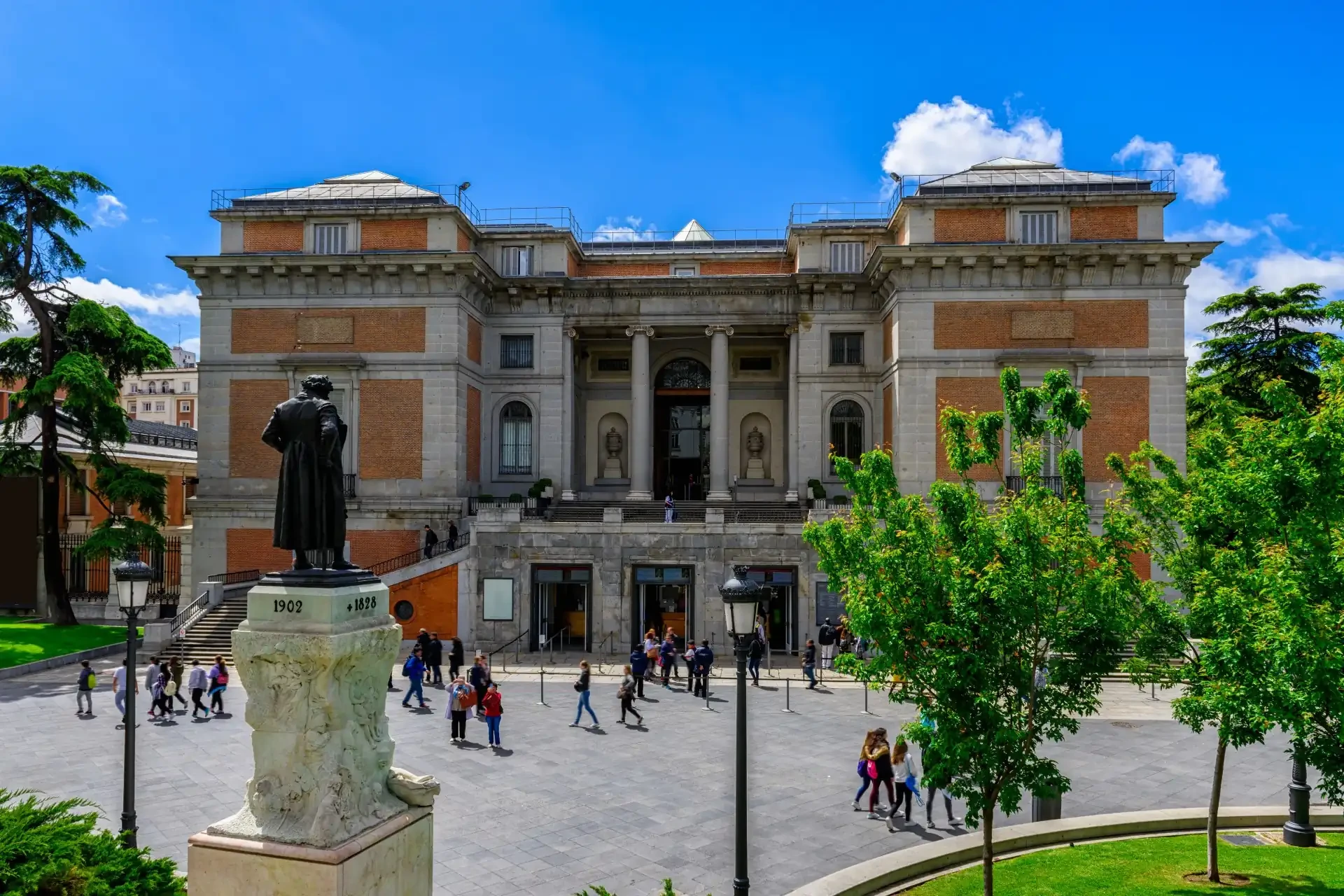 The Museo Nacional del Prado, also known as the Prado Museum, located in Madrid, Spain, is shown with people walking outside, set against a backdrop of cloudy blue skies.