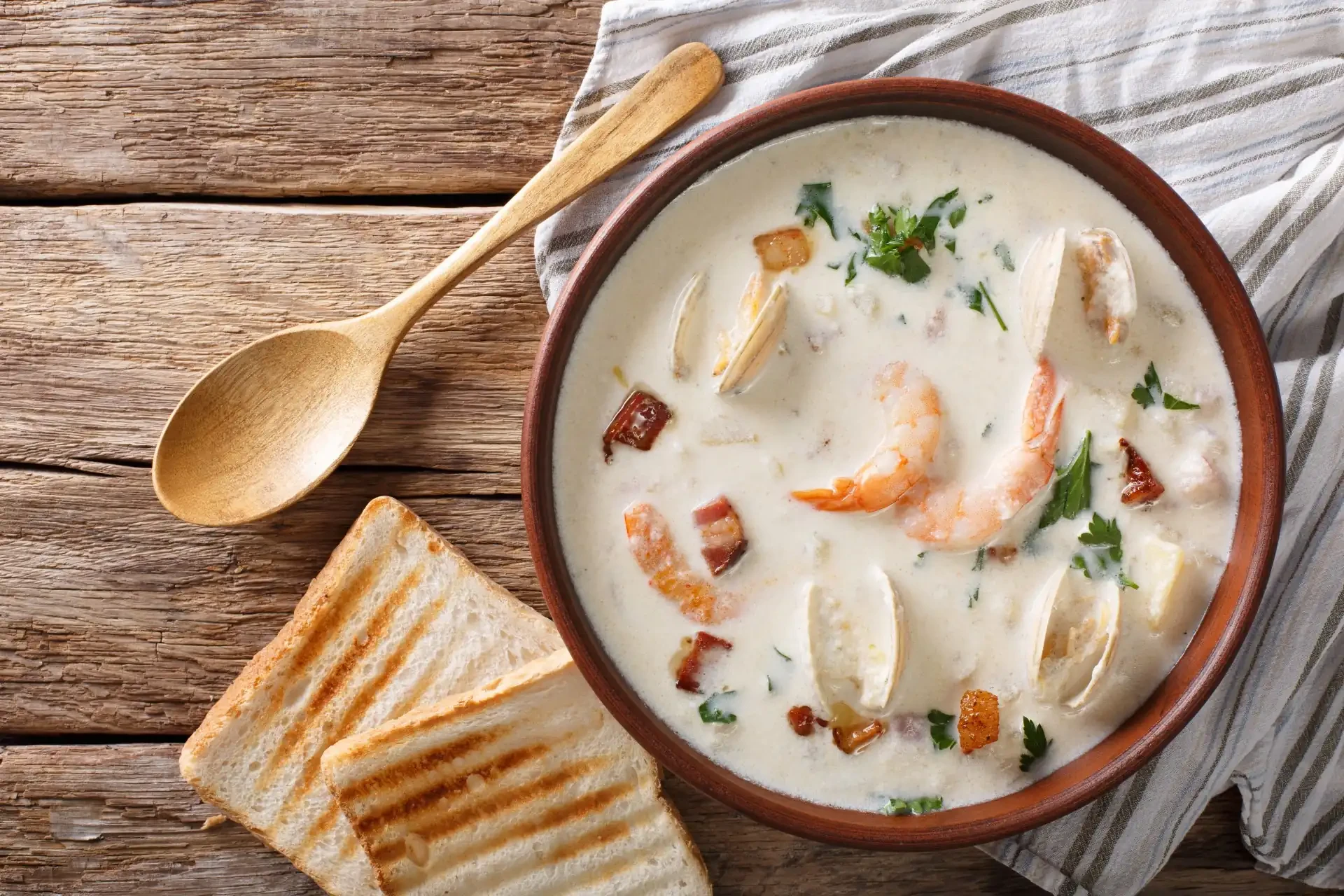A close-up of a bowl filled with New England clam chowder, featuring shellfish, bacon, and parsley, with bread on the side.