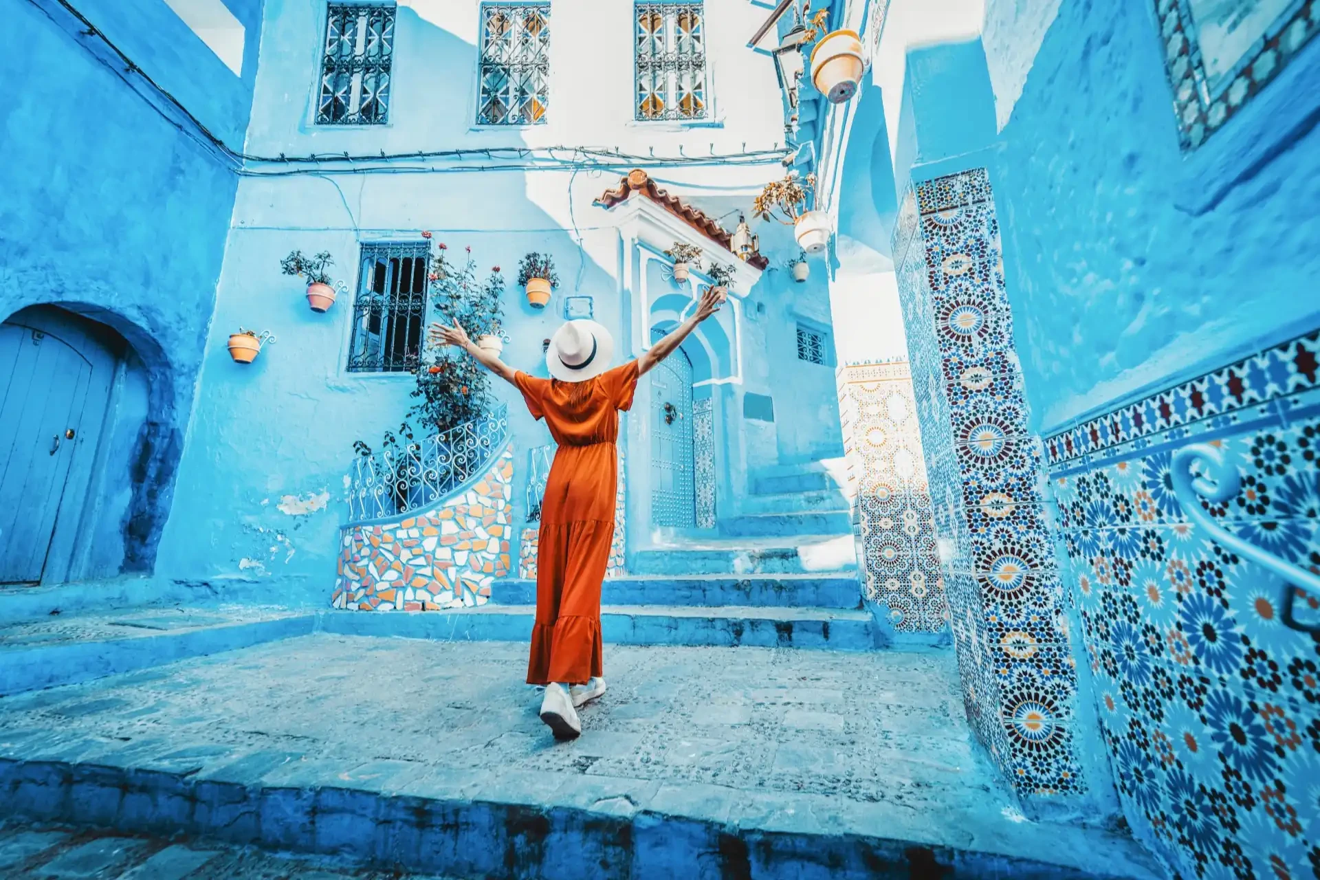 A woman in an orange jumpsuit and white hat walks through the streets of Chefchaouen, Morocco.