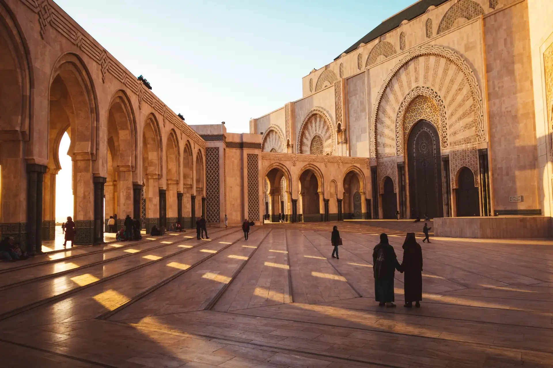People walking through the grounds of the Hassan II Mosque, Morocco against a blue sky backdrop.
