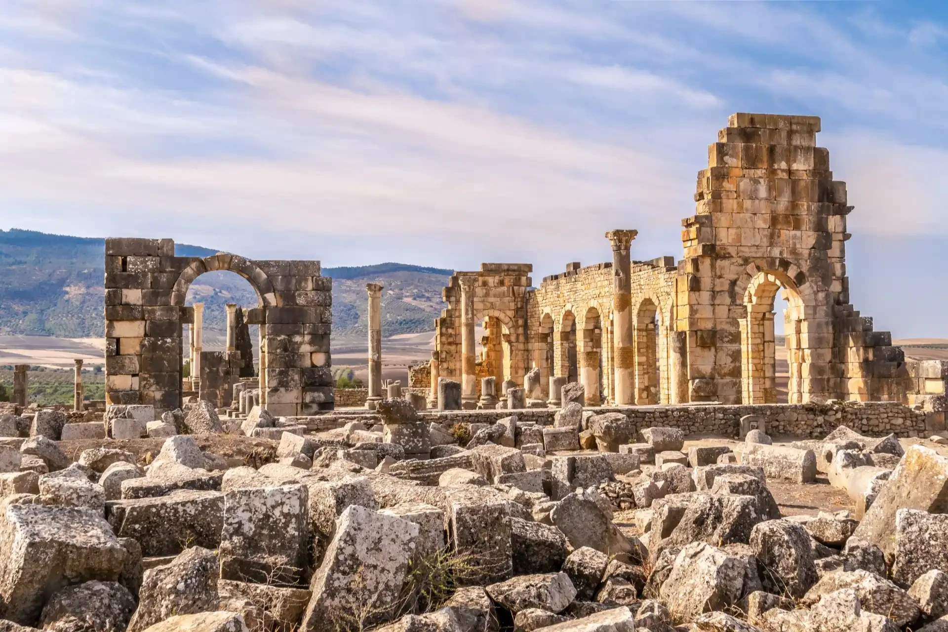 Ruins of Roman structures surrounded by mountains under a cloudy sky.