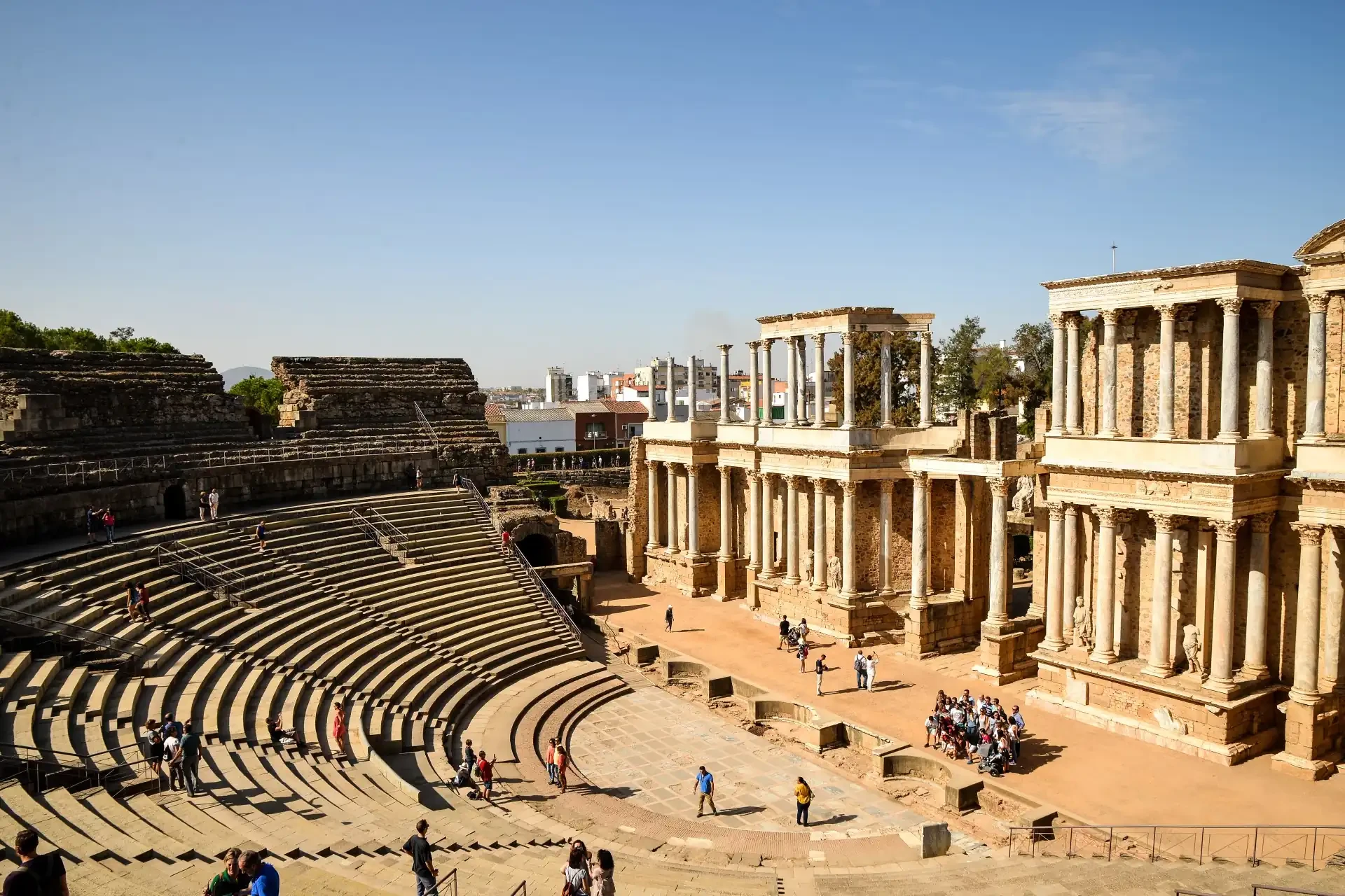 Ruins of an ancient theatre in Merida, Spain, are depicted with people walking around, possibly part of a tour group, exploring the historical site.