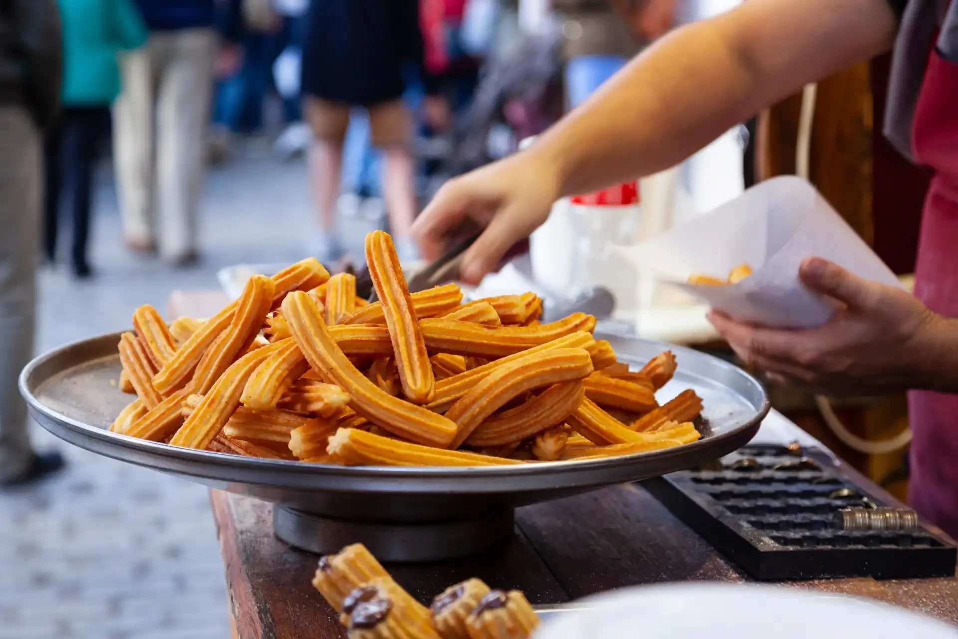 A person is seen serving freshly made churros with chocolate sauce in paper cones. The golden-brown churros, crispy on the outside and soft on the inside, are a popular Spanish treat enjoyed especially with a generous drizzle of chocolate sauce.