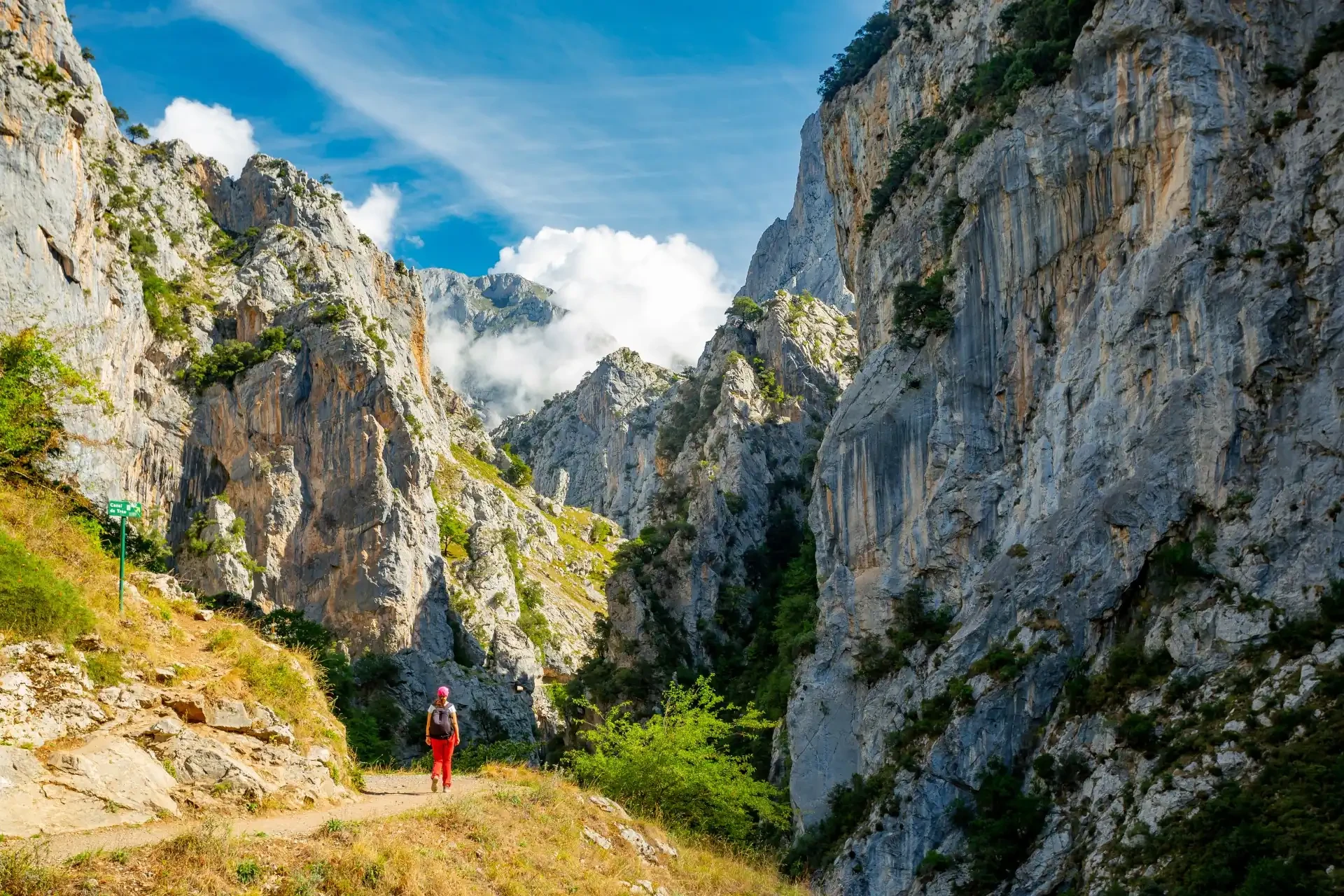 A lone hiker wearing red trousers, a light-coloured t-shirt and a pink cap is seen trekking along the Ruta del Cares trail in Picos de Europa National Park, Spain, with towering mountains, blue skies, and wisps of clouds creating a picturesque backdrop.