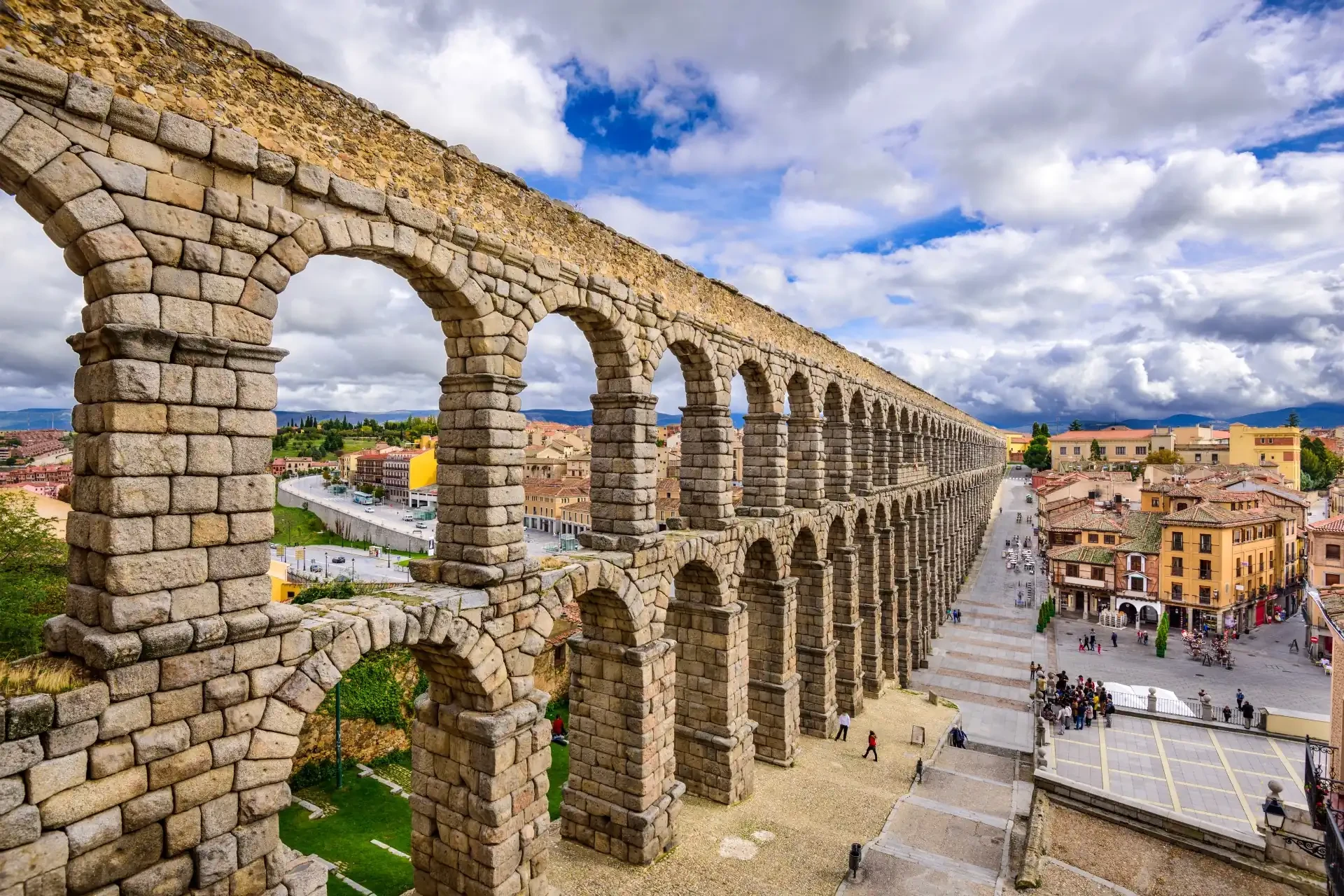 The Segovia Aqueduct, featuring its iconic arches, is showcased against a cloudy sky backdrop, with colourful buildings and pedestrians adding to the lively atmosphere of the scene.